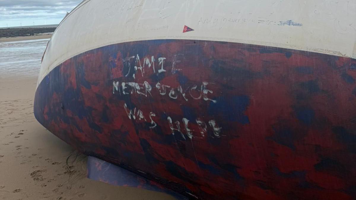 White writing is on a red and blue patch of a yacht, which is sitting on its side on sand.