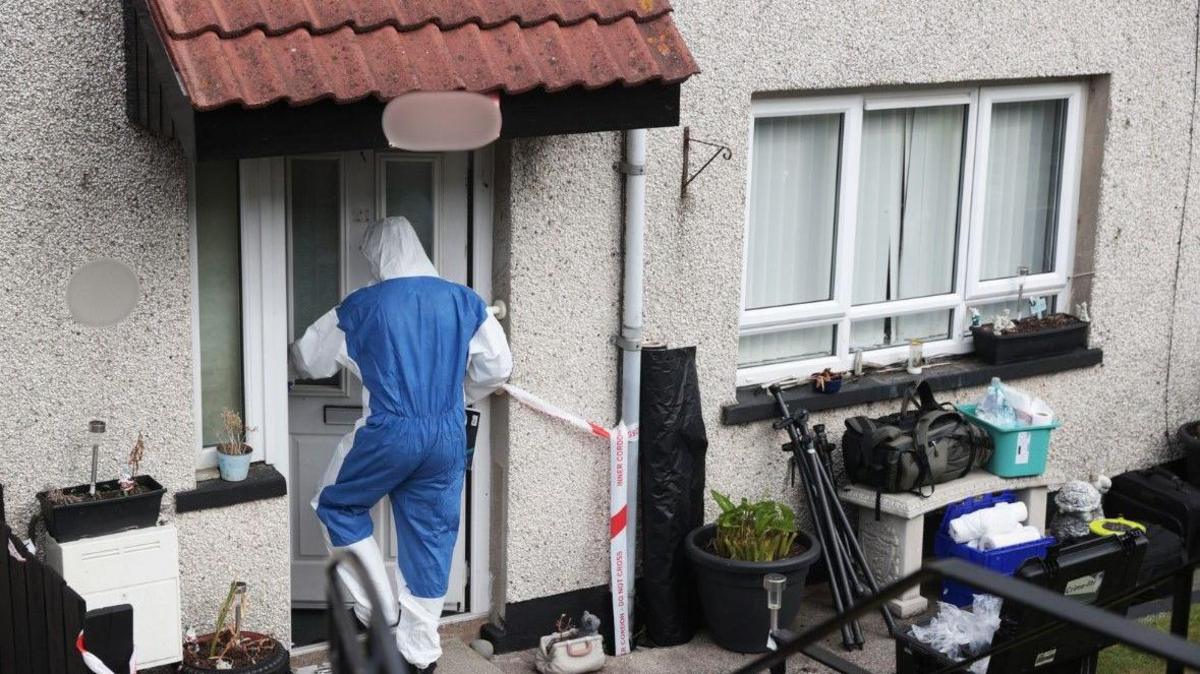 An individual in a forensic protective suit is walking into a property