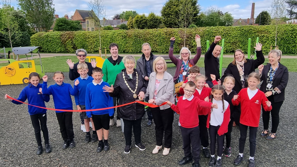 Schoolchildren and a group of adults cheer as a red ribbon is cut in a play area