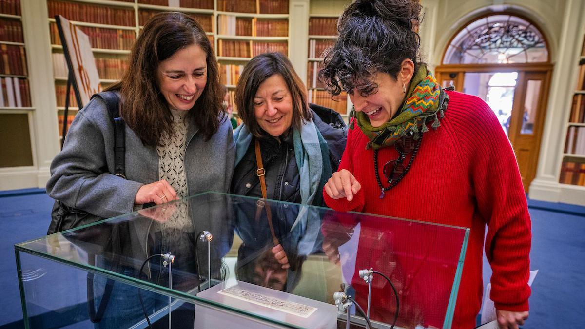 Three woman looking at the parchment, which is sealed in a glass case. The first woman is wearing grey jacket and has brown hair, with a black bag over her shoulder. The middle woman has brown/black hair and a grey jacket, while the woman on the right of the picture has a red jumper and green scarf on, with black hair. Bookcases are behind them.