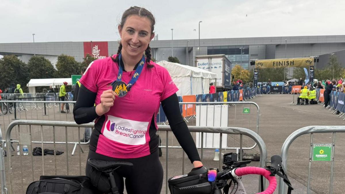 Victoria Coles holds a bicycle near a large finish banner. She wears black and pink cycling kit. Her hair is in braided pigtails.