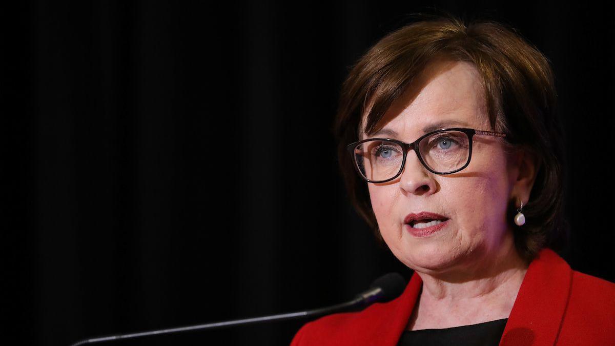 A woman with dark brown hair is talking at a podium. She is wearing a red blazer and dark rimmed glasses. 