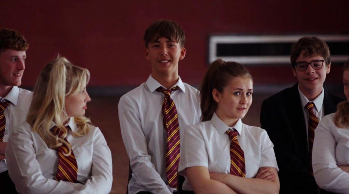 Dexter Henson is wearing a shirt and tie in a school uniform, surrounded by several other teenagers wearing the same. He is sat upright, smiling