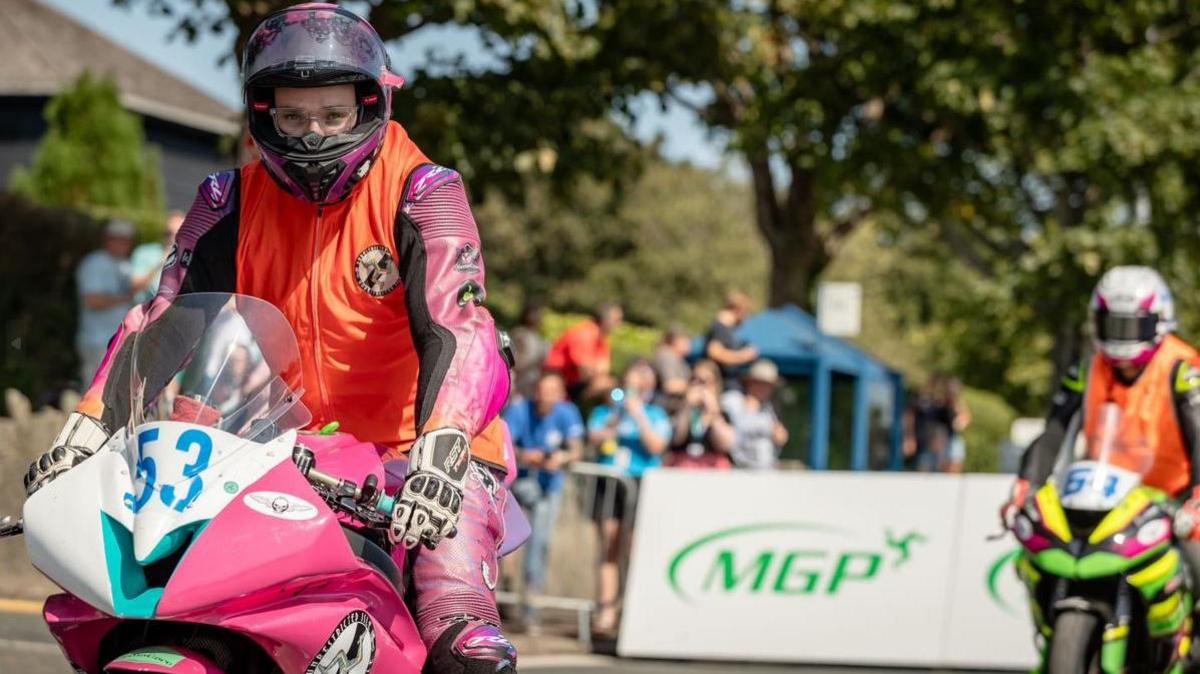 Blake Kelly wearing an orange bib over pink leathers and sitting on his stationary pink, white and blue bike. The visor of his helmet is up and his eyes are focused on the road ahead. There are large white boards with the green MGP logo on them in the background along with another rider in an orange bib on a green and yellow bike and with a group of spectators watching on.