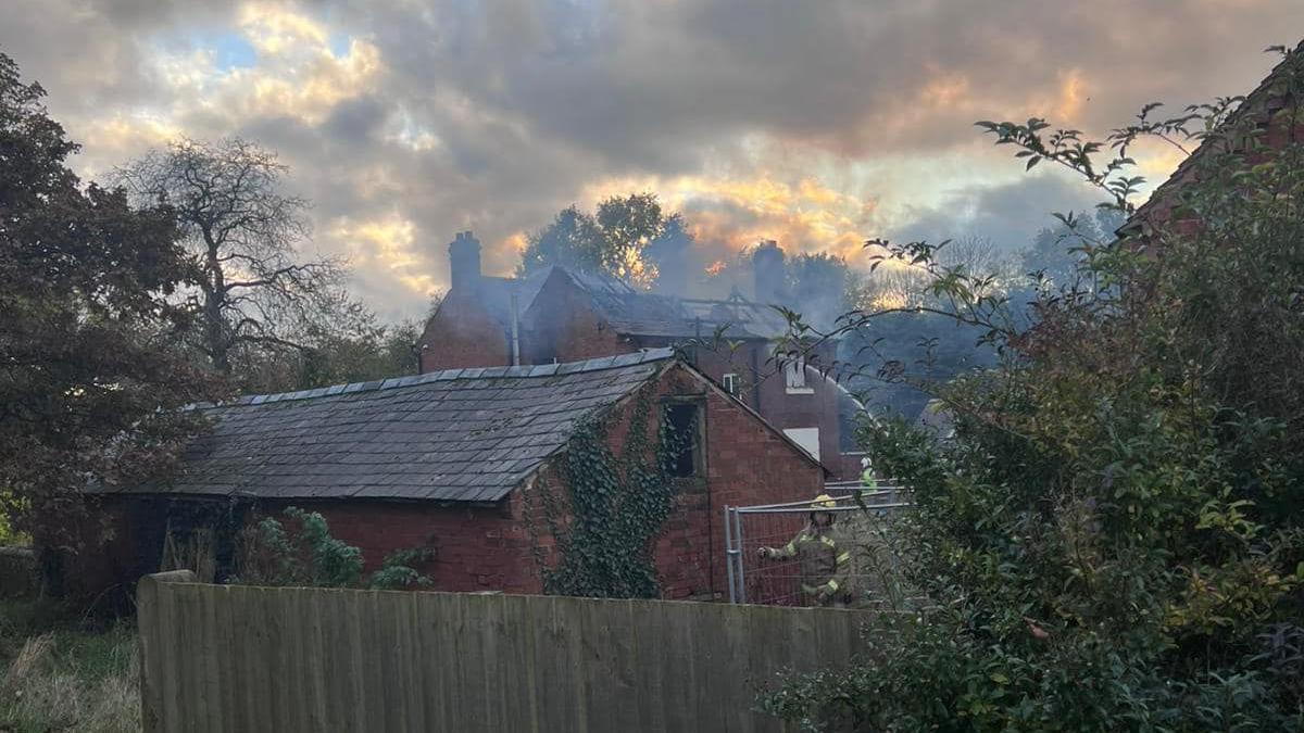 The charred roof of a building can be seen with smoke around it, as firefighters on the ground are directing water in through a second floor window.