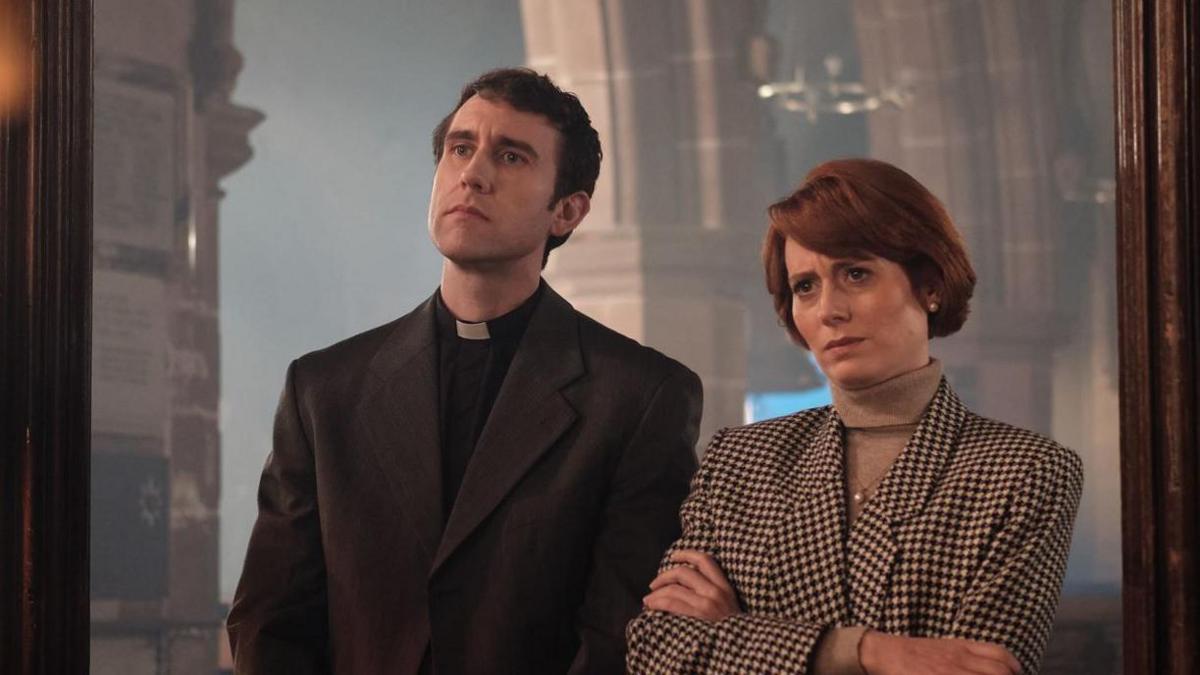A man with dark hair and black vicar's clothes alongside a woman with red hair in a chequed black and white jacket standing in a church