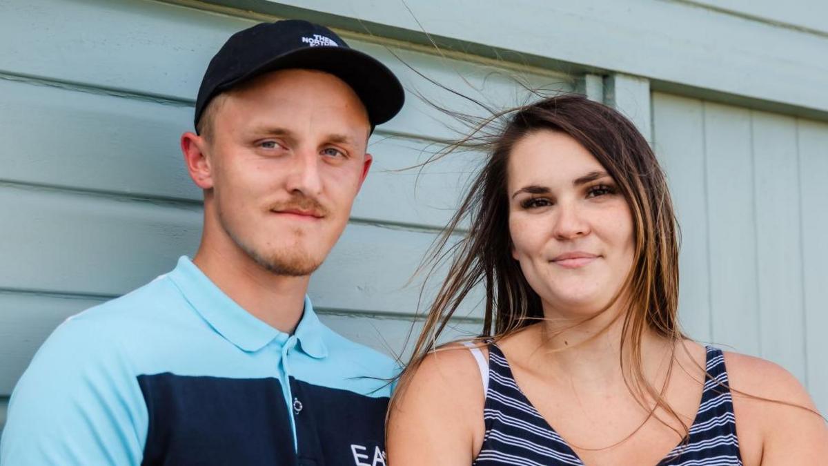 Matthew Steward wearing a black cap and blue polo shirt. He is standing next to Dannielle Rose, who has brown hair and is wearing a striped vest top. Both are in front of a teal beach hut and smiling.