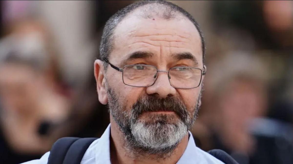 Andrew Malkinson with grey hair and beard wearing glasses and a light blue shirt with a ruck sack on his back.