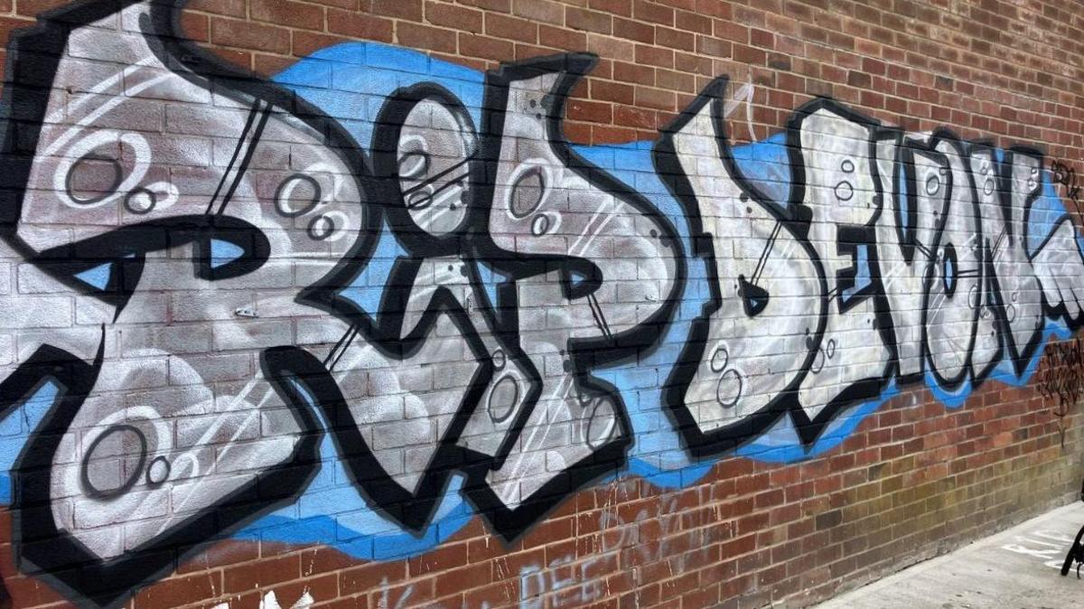 "RIP Devon" has been spray painted on to the brick wall of a building in blue, silver and black paint.