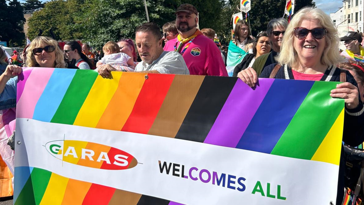 Adele Owen in a crowd of GARAS clients and supporters on a sunny day, holding up a rainbow-coloured banner saying GARAS welcomes all