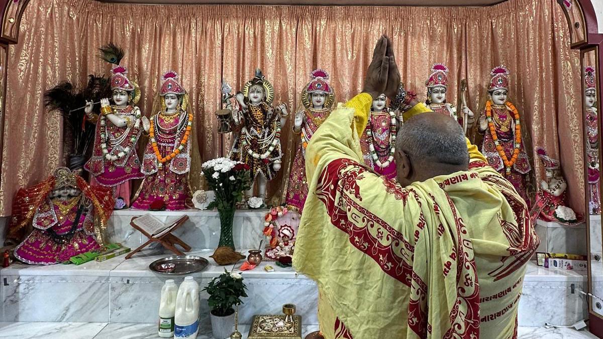 A Hindu priest prays to deities in the Main Sanctum of the Hindu Temple. He has his hands together above his head in prayer. He is draped in a yellow and red robes.