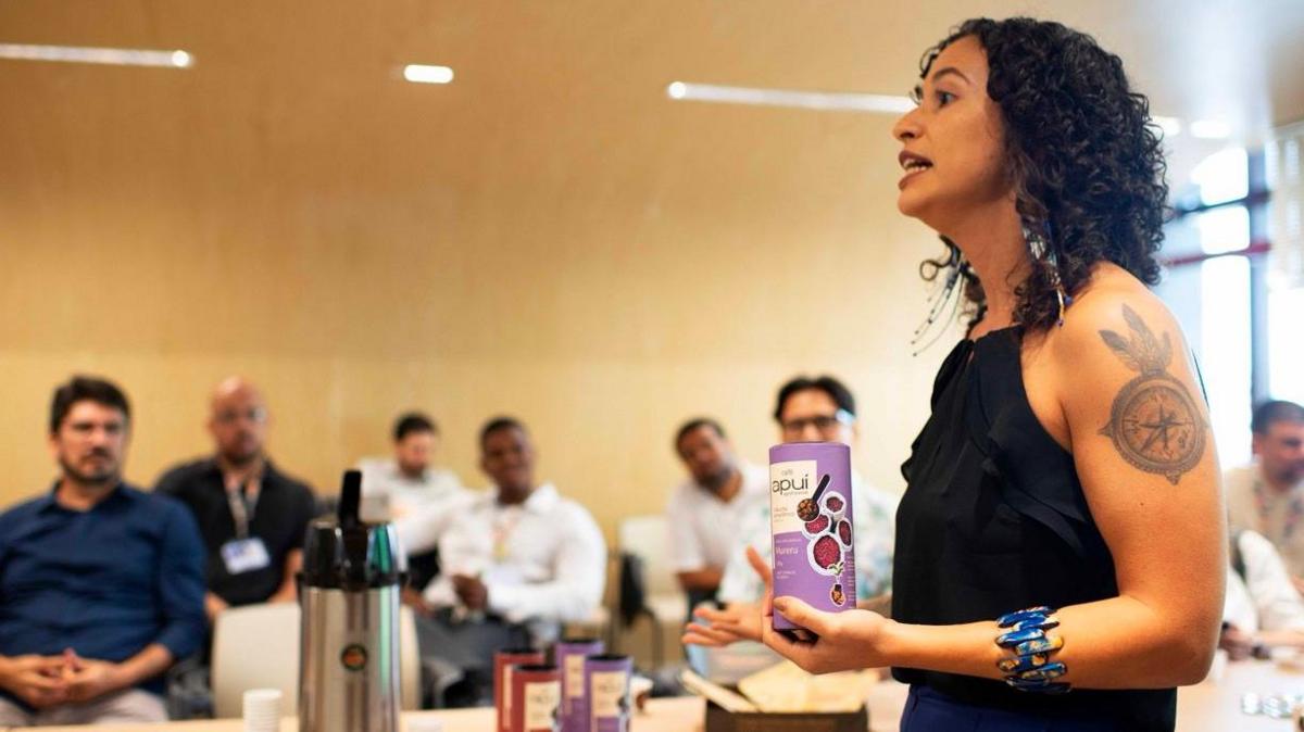 A woman stands in an office space holding a small purple tube with pictures of coffee beans on the side. She is talking. A group of men are in the background sitting down to listen to her.