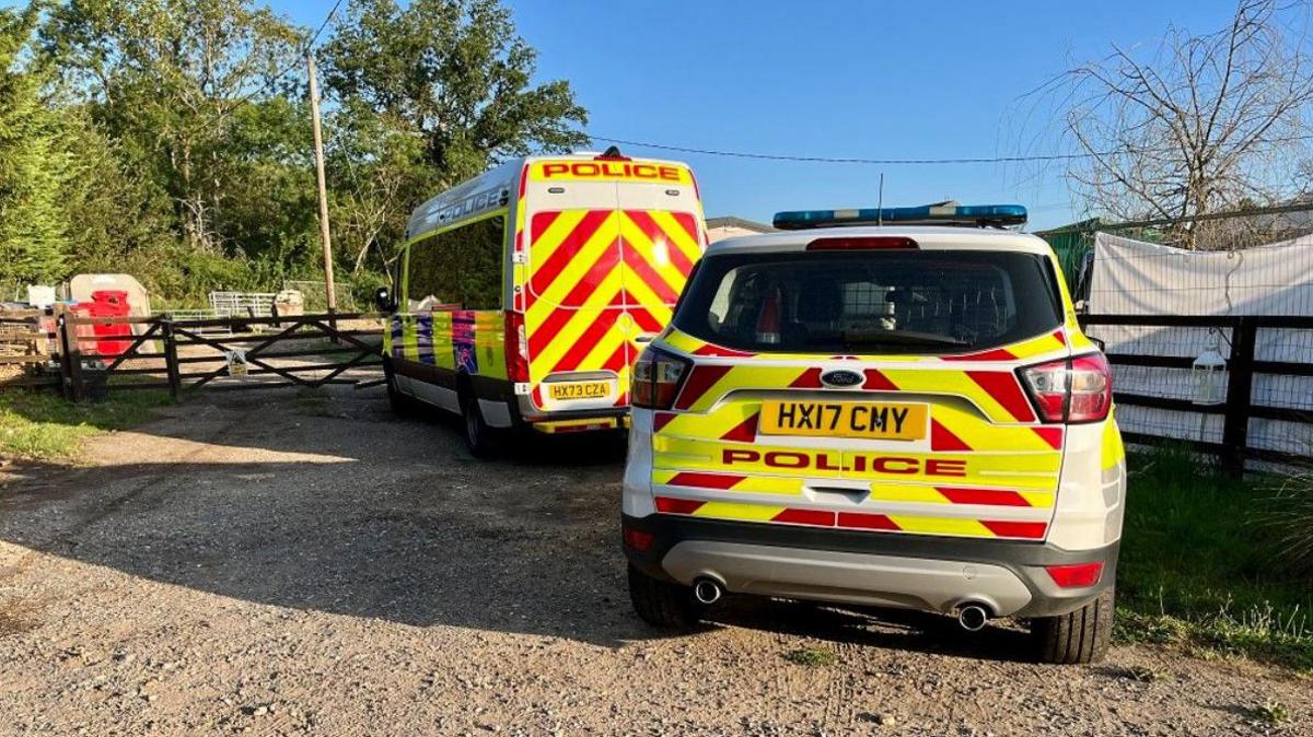 A police van and a police car parked next to a closed five-bar gate.