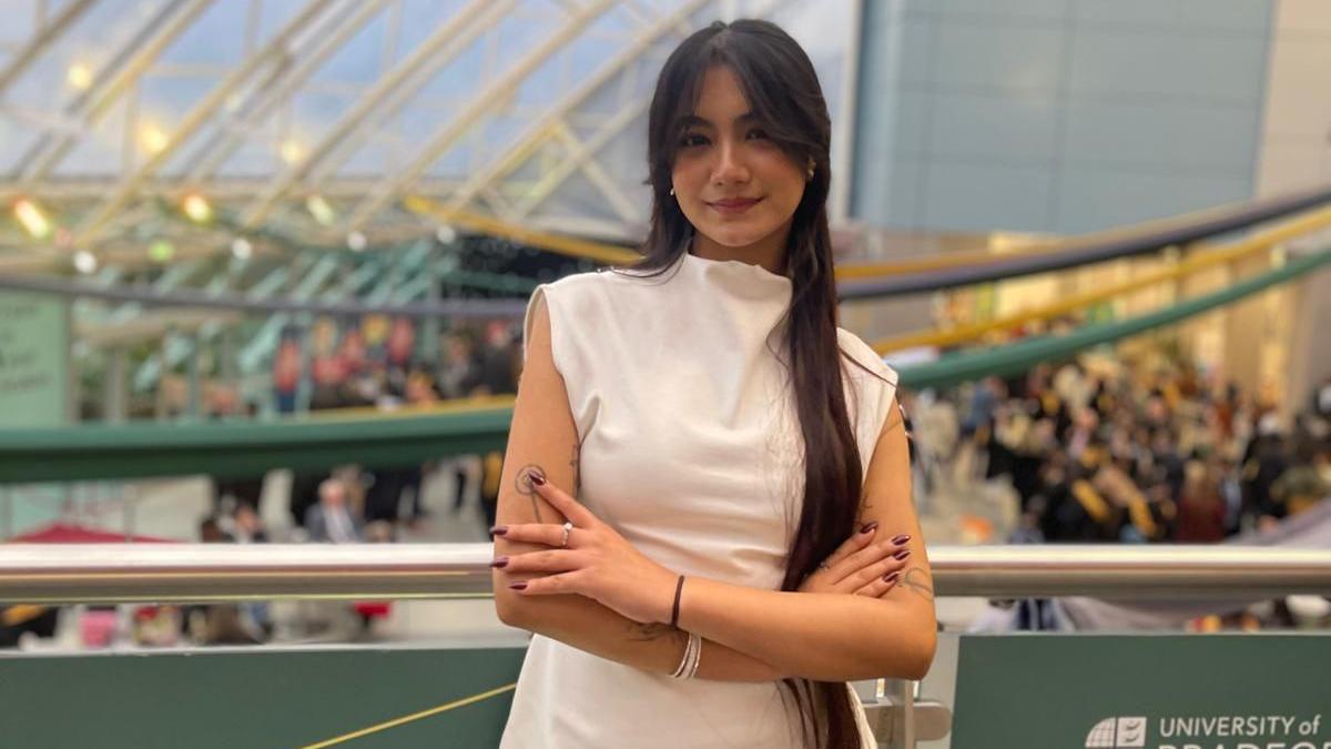 Sanskrity is standing in front of a busy hall. She is wearing a white dress and is looking determinedly at the camera