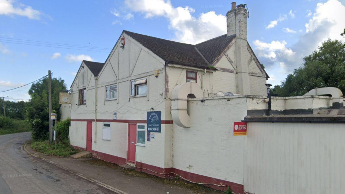 A screenshot from Google street view showing the White Horse Chinese restaurant as seen from the road. It is an old white building with red doors and a tall chimney. It is situated next to the pavement, surrounded by trees. There is a red sign on the building advertising the restaurant for delivery takeaways.
