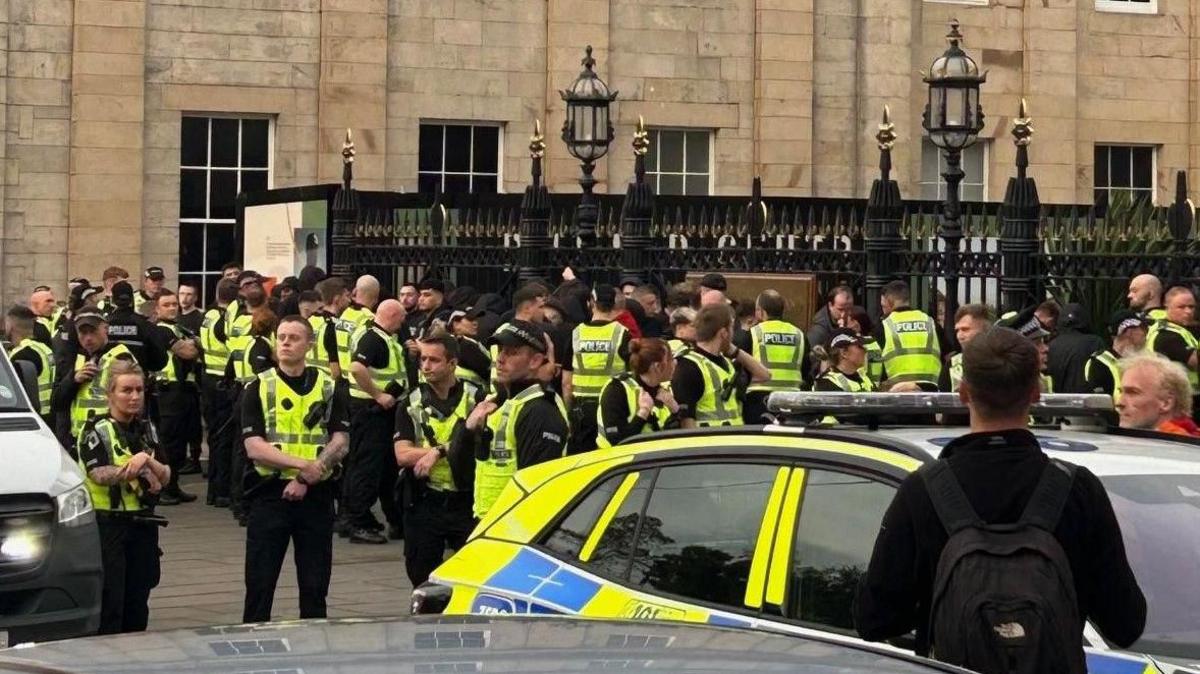 Several tens of police officers attend disorder in St Andrews Square, Edinburgh. They are standing on the pavement between a black gate and parked cars.
