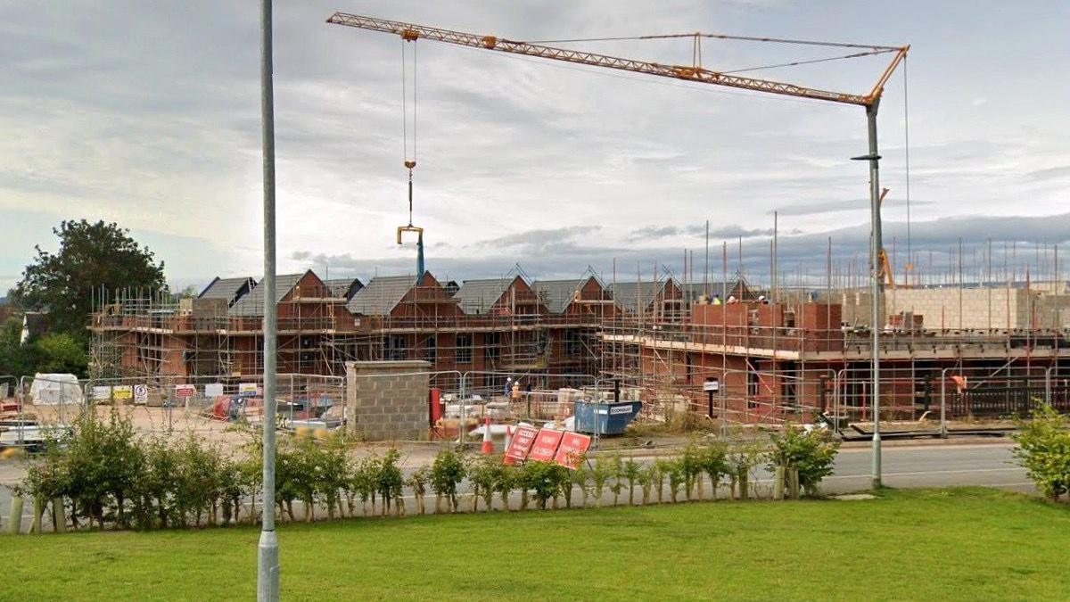 A street view of a construction site where homes are being built in Herefordshire. A crane towers over a row of houses that are being built. Road signs block the entrance to the site.