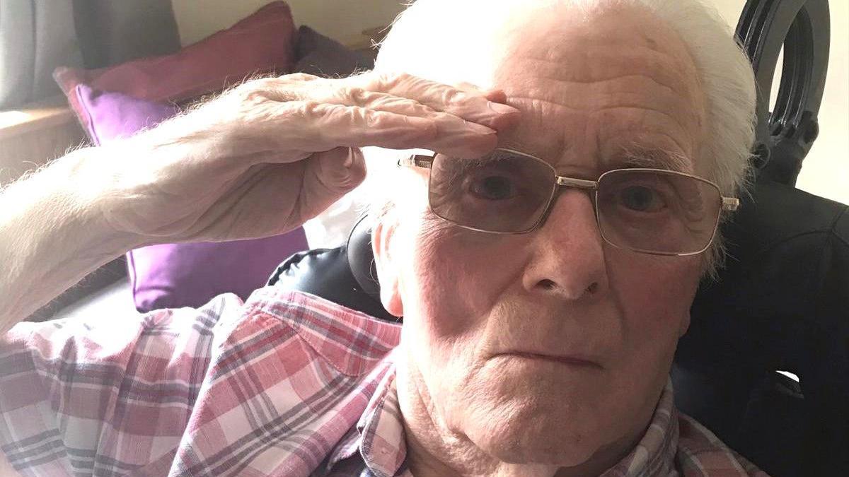 Maurice Page is pictured saluting to the camera. He is sitting on a chair wearing a light red, black and white chequered shirt with tinted glasses with silver coloured frames.