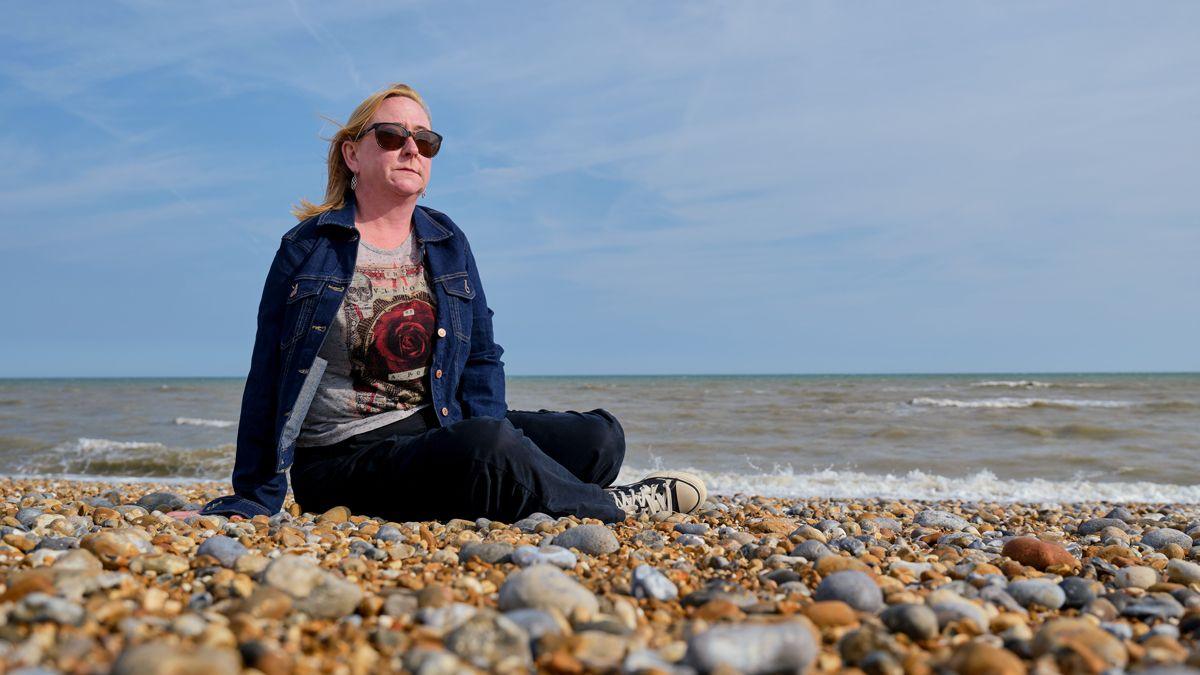 A woman with blond shoulder-length hair, wearing sunglasses, who is sitting on a pebble beach close to the water's edge. The sea is behind her and the sky is blue. She is wearing a blue denim jacket and a top with a big red rose on it. 