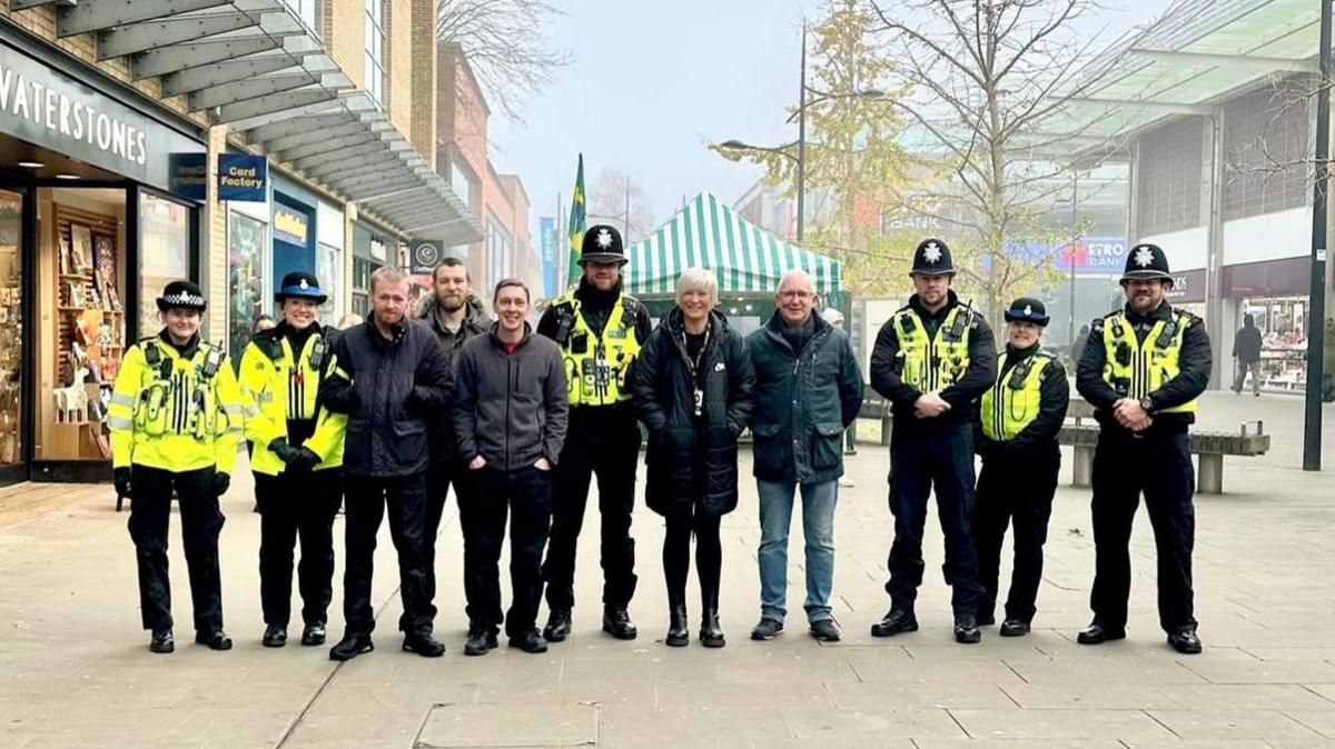 The neighbourhood policing team stand in a line in the town centre looking at the camera. Six people are wearing full uniform, whereas five people are in plain clothes.