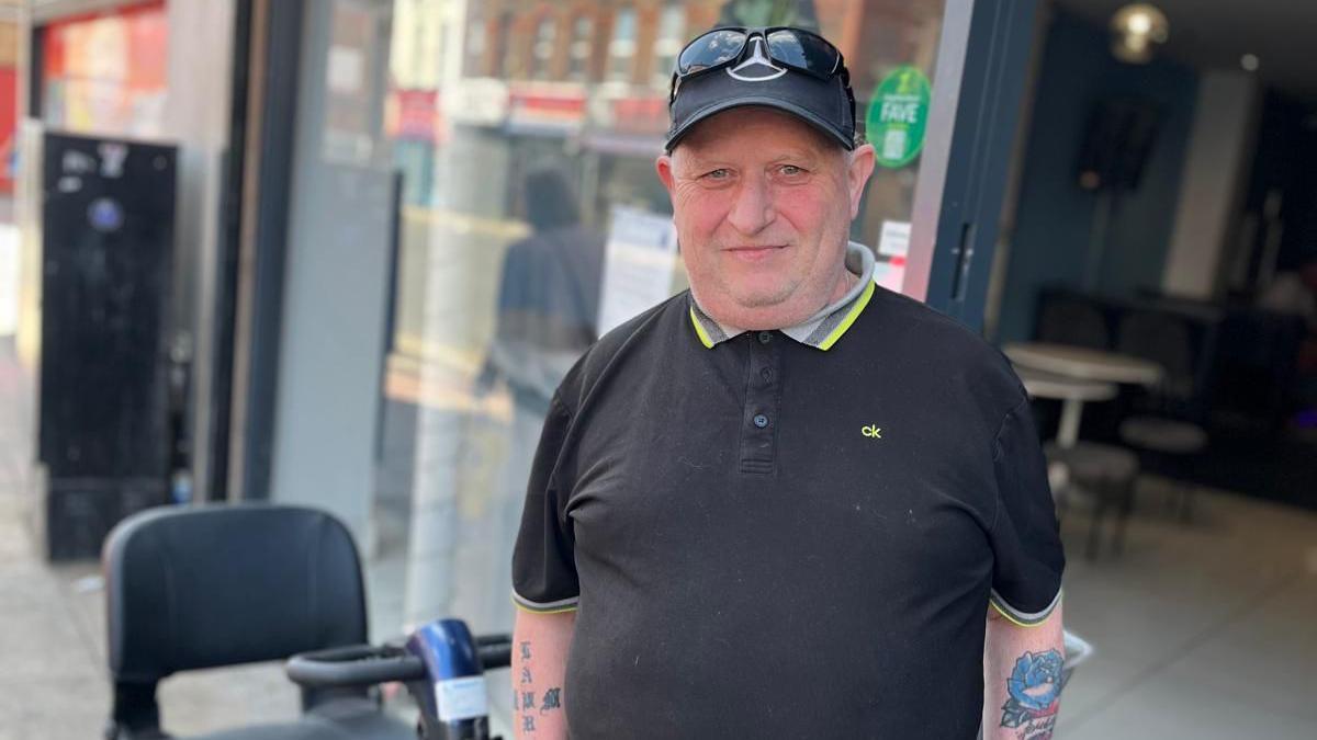 Jimmy Williams, wearing a black polo-neck t-shirt, black baseball cap and sunglasses on his head, smiles at the camera on the street at County Road.