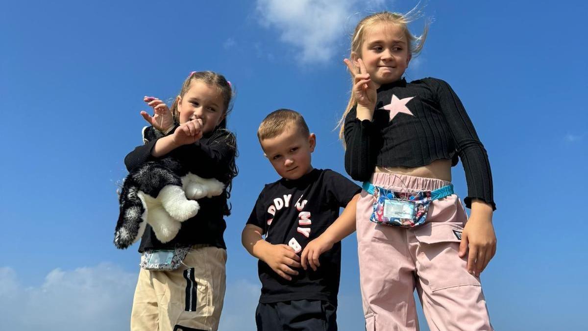 Siblings Paisley, Louis and Evie are standing side-by-side on a summer's day. The camera is pointing upwards, meaning that we don't see the ground, only the blue skies behind them. Paisley has her arms crossed in the air and is holding a cuddly toy. Evie is making a peace sign. Louis in the middle is staring directly at the camera. 