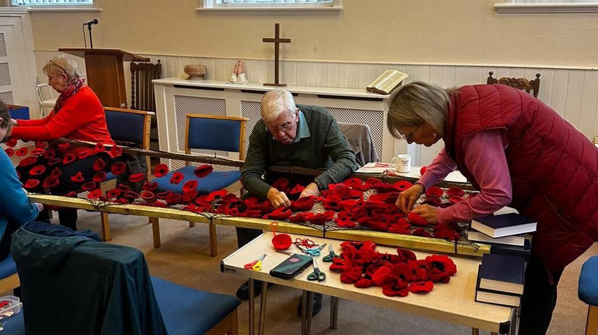 An elderly man and a woman in a red coat hand sewing multiple knitted poppy flowers onto a netted mesh