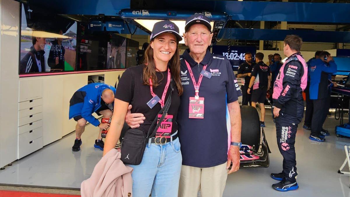 And older man and young woman wearing VIP passes stand in front of a Formula 1 garage labelled. Team members are visible inside working on equipment and cars.