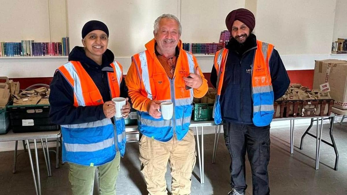 Kully Kaur Deol, Marcus Lapsa and Bill Mato are standing in front of a table laden with sandwich meal bags and boxes of crisps in the Trallwn Community Centre in Pontypridd. They are all smiling and Kully Kaur Deol and Marcus Lapsa are holding cups, with Marcus Lapsa holding what looks like a mobile phone.