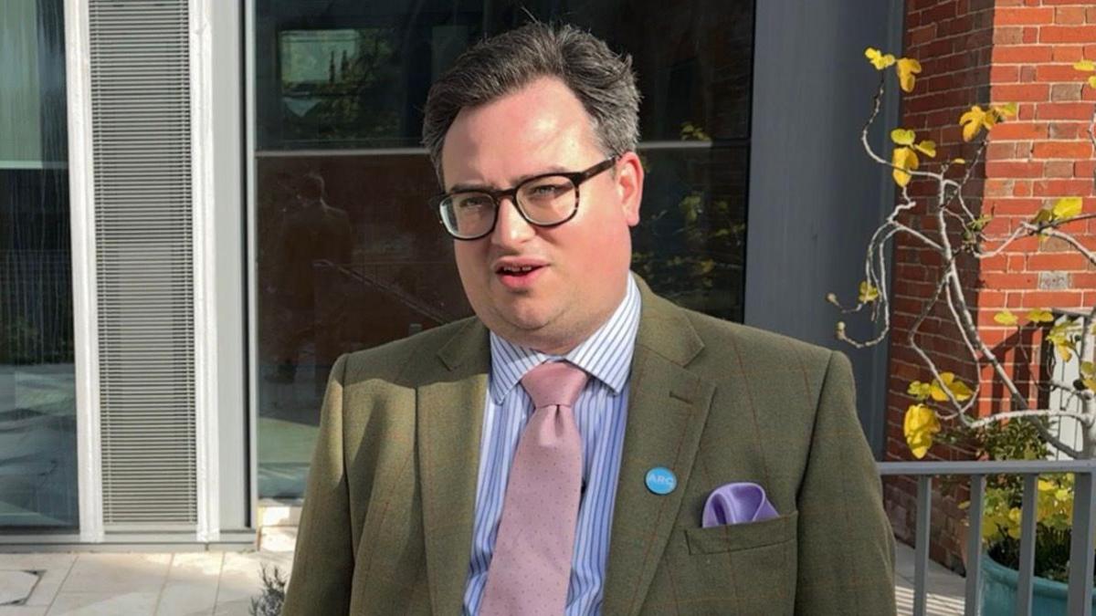 Ed Rimmer is wearing a blue and white shirt with a pink tie and green tweed jacket. He is standing in a stone paved garden with a flower pot and Wiltshire Council building in Salisbury behind him