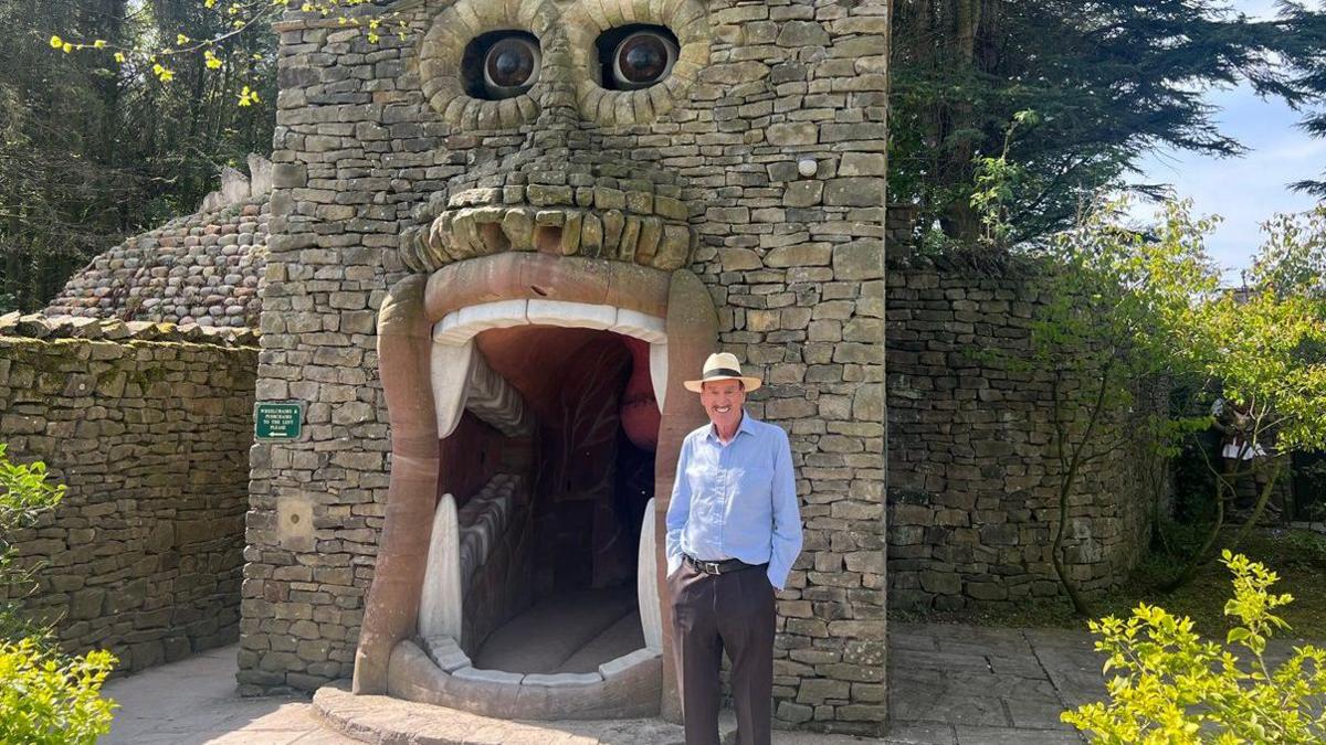 An elderly man in a blue shirt and cowboy-style hat. He is smiling widely and has a moustache. He stands in front of a stone doorway in the shape of a giant mouth with eyes above and big teeth.