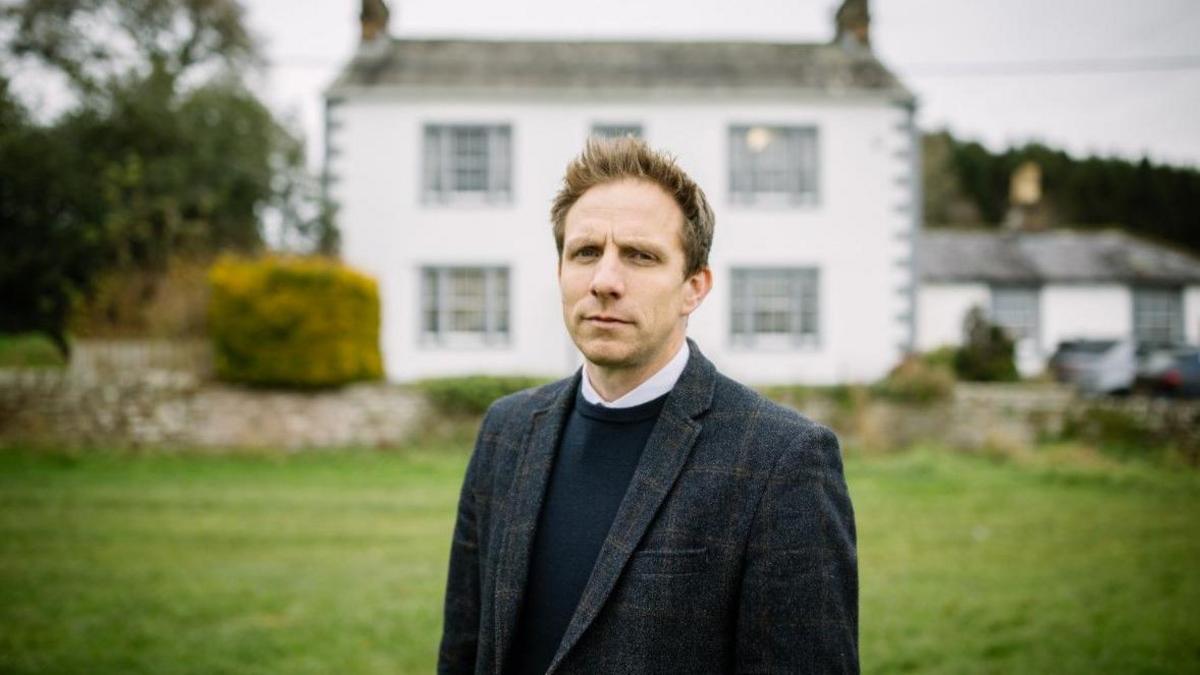 Paul Borrows standing in a grassed area at the front of Hunter Hall school. He has dark hair, spiked at the front, and is wearing a dark blazer and navy jumper.