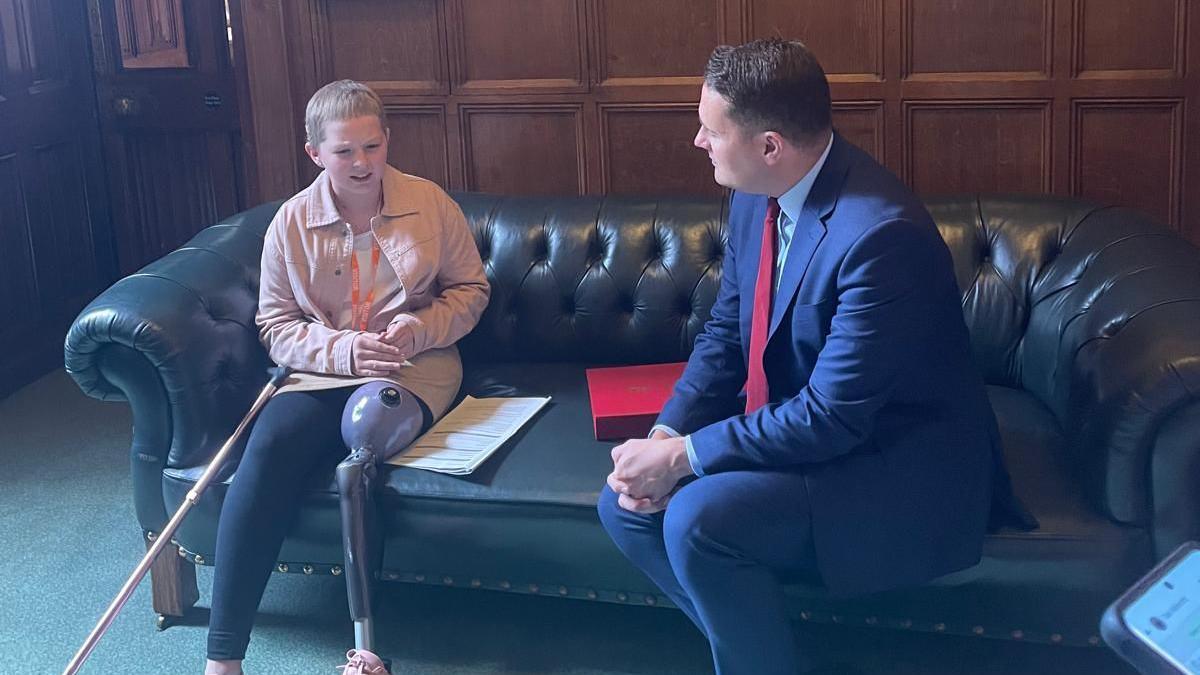 Millie, sitting on a large leather hair, with a false leg, next to Wes Streeting, who is looking at her. They are both mid conversation. A wooden panelled wall is behind them.
