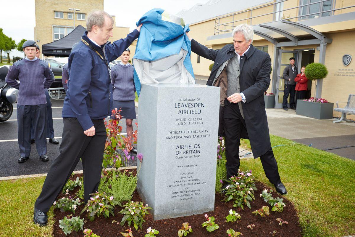 Kenneth Bannerman is wearing black trousers, black shoes and a navy coat and lifting a cover off of a grey granite stone with the help of another man. The stone is in the shape of a cuboid and its front reads "In Memory of Leavesden Airfield". Members of the RAF watch on in the background. To the right is the Warner Bros logo next to the entrance to the studio.