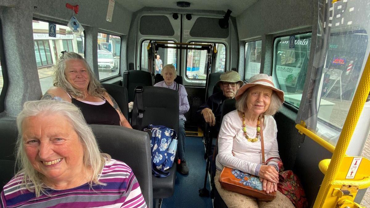 Five elderly people in a minibus, looking at the camera and smiling