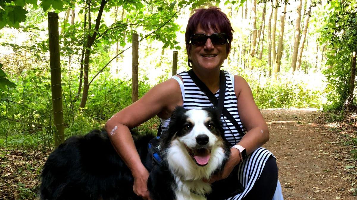 Cheryl squats next to her dog Freddie in woodland, with sun streaming through the trees. She is tanned and has cropped burgundy coloured hair, and wears sunglasses and a striped sleeveless dress. She smiles at the camera and has her arms around Freddie, a large black and white dog who appears to be an Australian shepherd dog. His mouth is open and he looks at the camera.