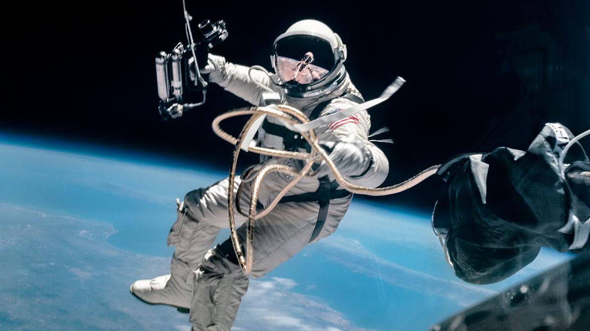 An astronaut in a white space suit holding onto a cord connected to a spacecraft and walking in space. The surface of the earth can be seen in the background. 
