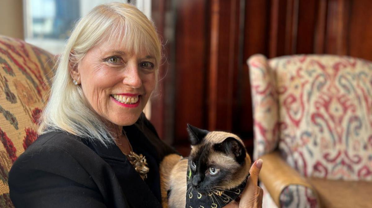 Holly Hennessy is smiling at the camera whilst holding her cat captain. Holly is wearing a black blazer and has shoulder-length blonde hair. Captain the cat is a light brown colour with black ears, a black stripe between his eyes and a black patch around his mouth.