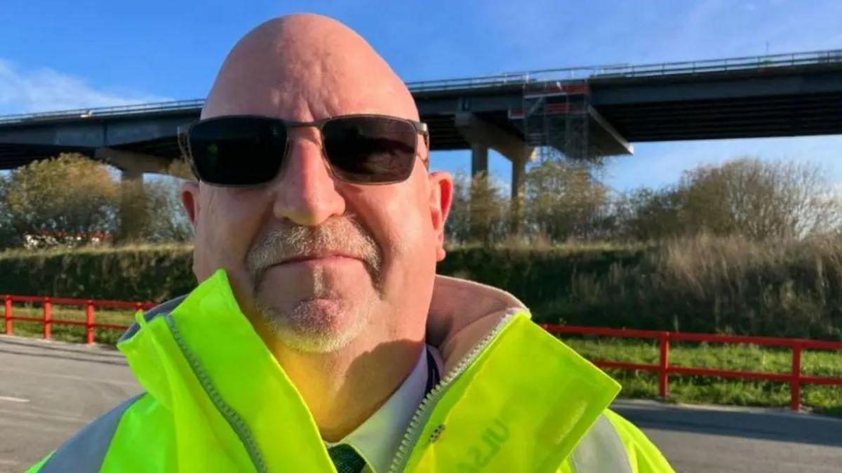 A bald man with glass sunglasses and a white moustache looking into the camera. He is wearing a high-vis yellow jacket over a white shirt and tie. There is a grass verge and a tall bridge behind him.
