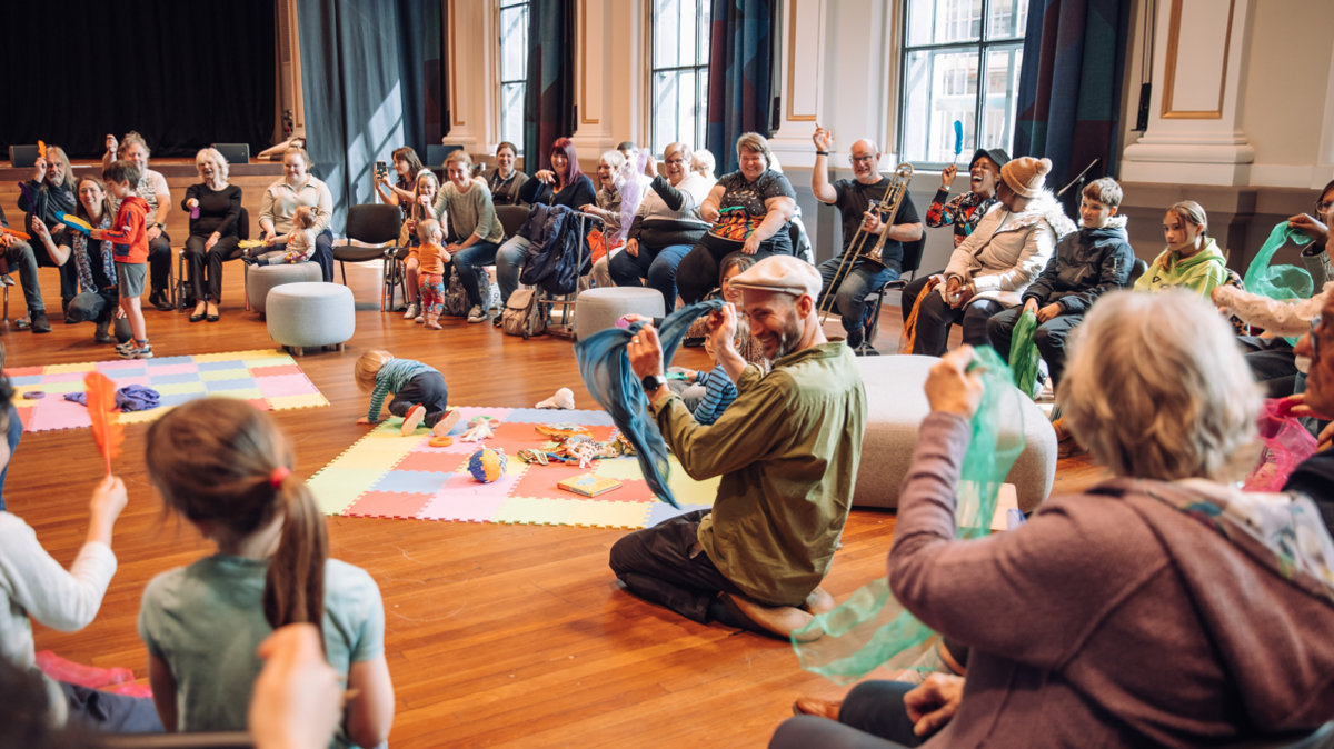 Lots of people sitting in a circle in a workshop at the Bristol Beacon. There are soft mats in the centre with children playing, people waving silk scarves and others with instruments. 