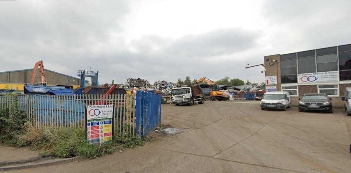 A scrapyard on a cloudy day. There is a car park and a two-storey office building on the right. A truck, diggers and piles of scrap metal can be seen in the background on the left.