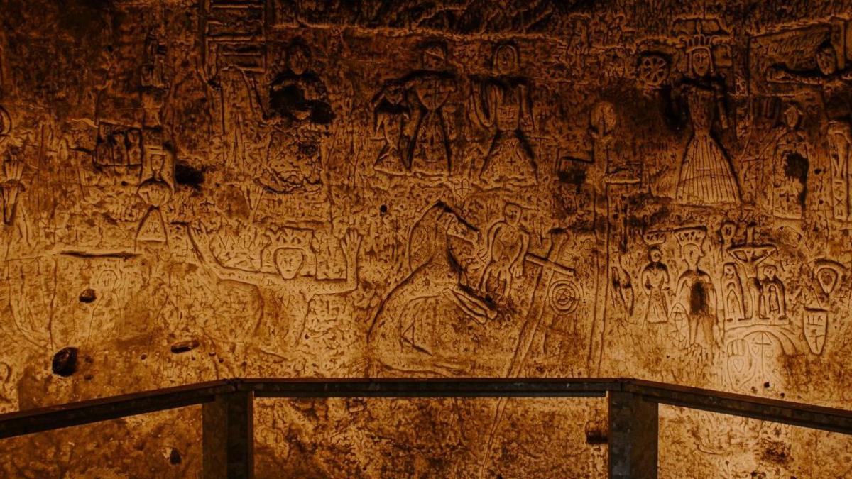 A lit up wall of the dark cave, showing many carvings of people and scenes. In the foreground is a wooden barrier.