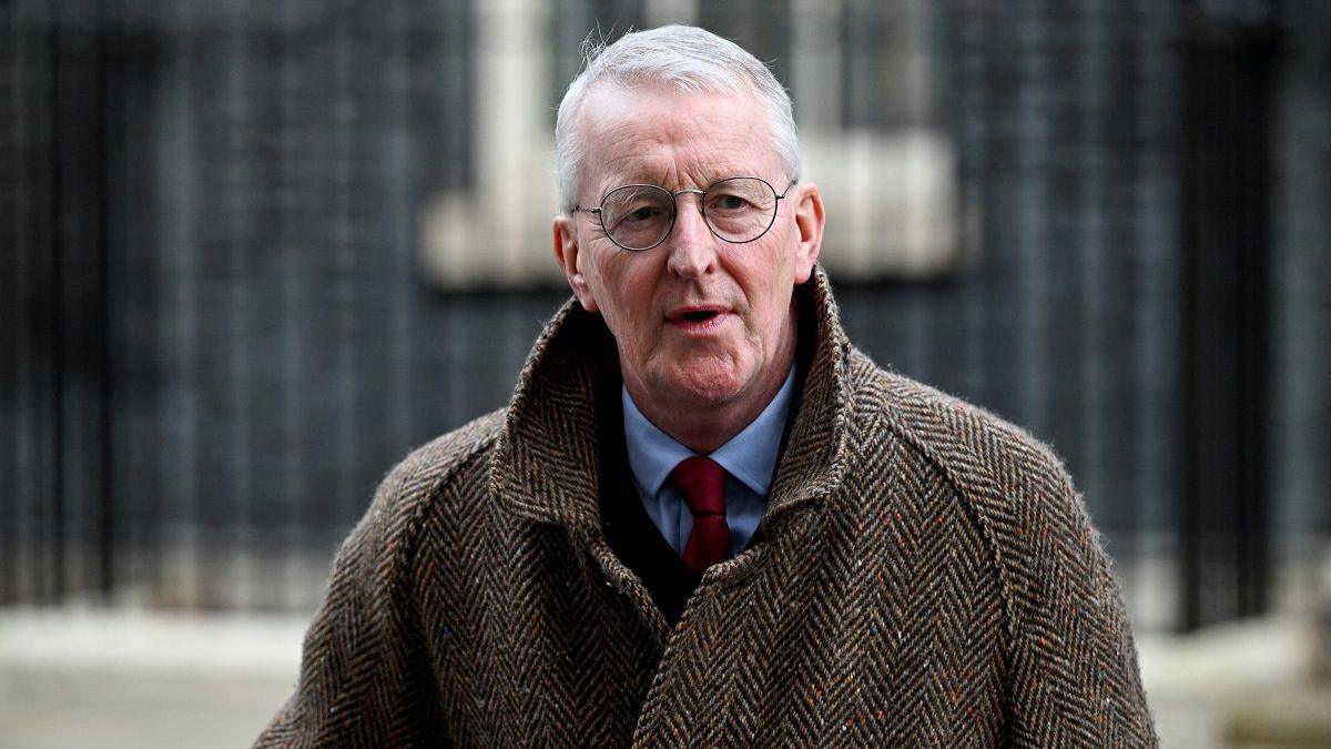 The Secretary of State for Northern Ireland, Hilary Benn MP in a black suit and red tie, with a tweed coat over it. He is mid speech. He has white hair and glasses