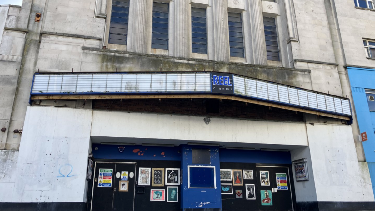 The image shows the exterior of a building that was once a REEL cinema. It has a marquee sign with the name, but the structure looks worn and possibly abandoned. The marquee itself is mostly empty, and the entrance appears to be boarded up, with framed posters or images displayed over the boards. The building has tall vertical windows and some decorative architectural features.