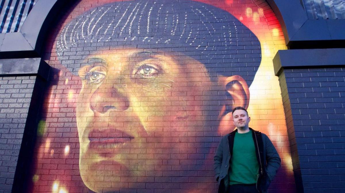 A man wearing a green jumper and a blue coat. He is standing in front of the mural of Thomas Shelby which has an orange and red painted background. Thomas is wearing a black flat-cap and staring into the distance in the mural.