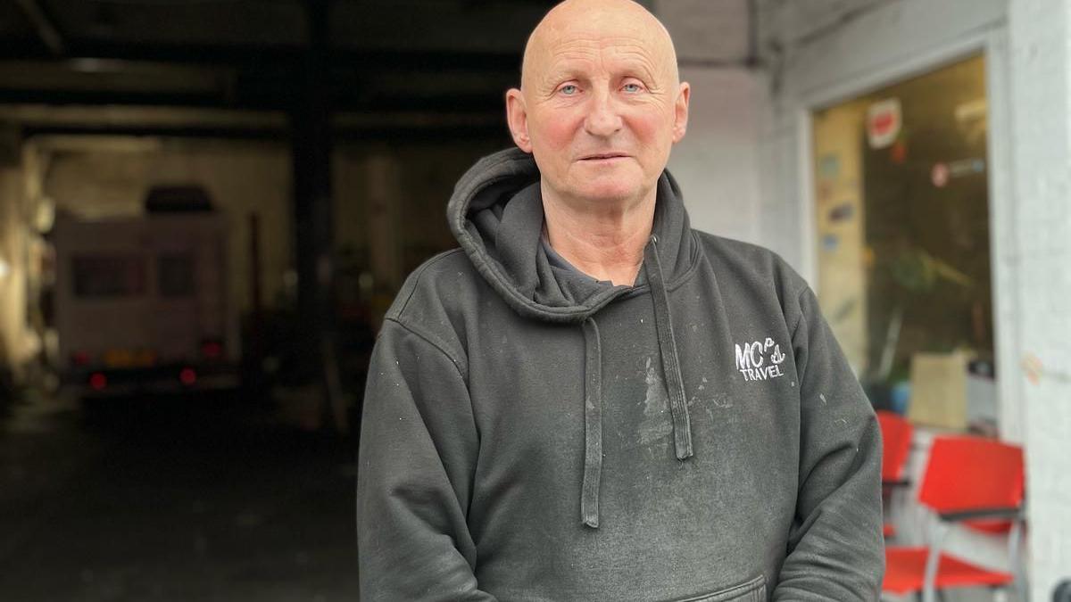 Paul Jones in his 60s, who has a bald head and blue eyes and is wearing a black hooded sweatshirt, looks into the camera with his garage workshop in the backdrop.