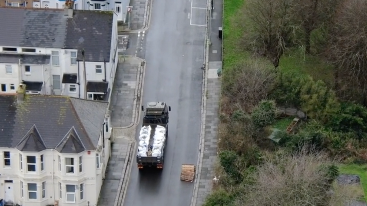 An aerial view of an army vehicle, with its back filled with sandbags and a WW2 bomb. The vehicle is driving up the empty street, there are no cards on the road. It is driving next to houses to the left of the road, and grass and tree to the right of the road.