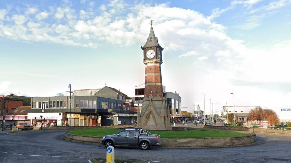 The Skegness Jubilee Clock Tower, which has a stone base, a red-brick column, four faces and a pointed bronze roof. It sits on a roundabout and a pick-up truck is passing in the foreground. In the background are the buildings of a seaside parade. 