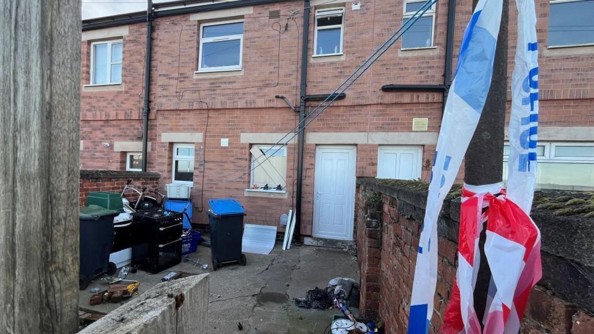 The exterior of a row of terraced houses. The patio out front is littered with appliances, including cookers, and police tape is tied to a pole in the foreground. No fire damage can be seen on the outside of the house but the bottom window is boarded up.