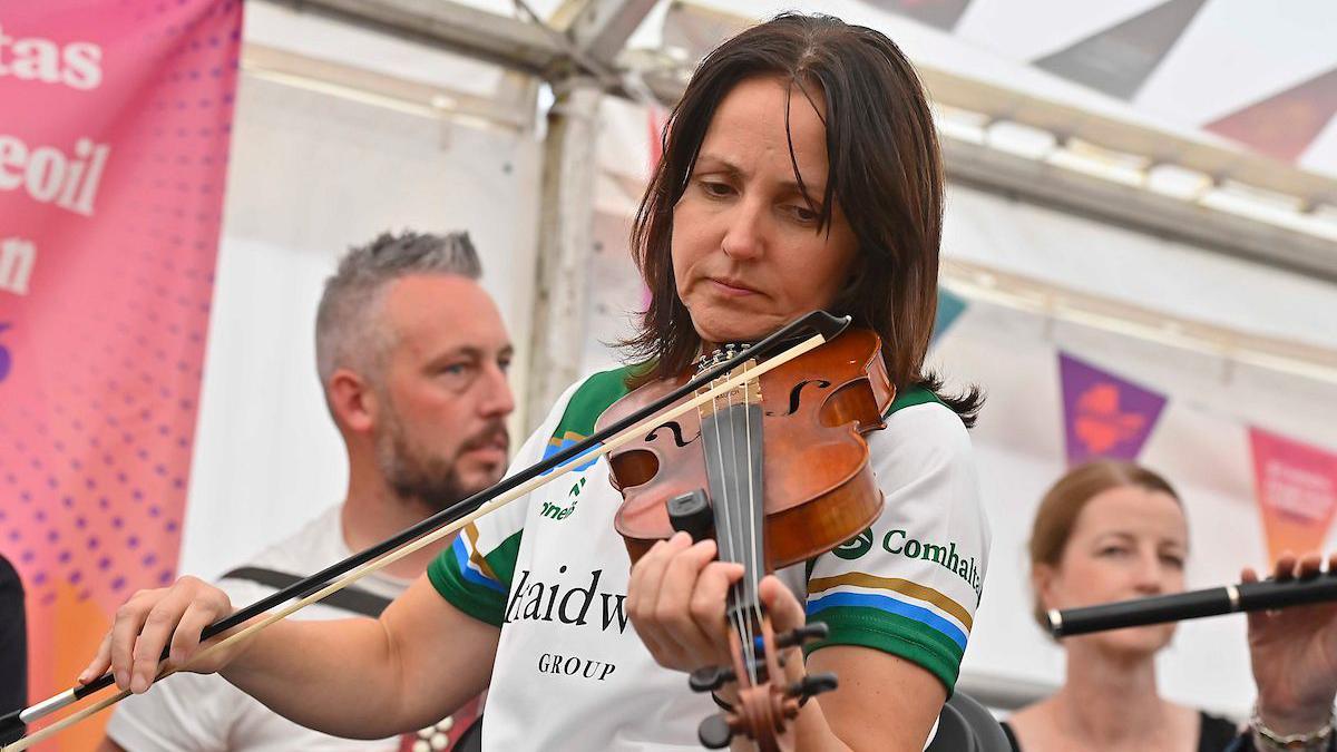 A woman with dark hair holding a fiddle. She appears to be standing inside a tent. 